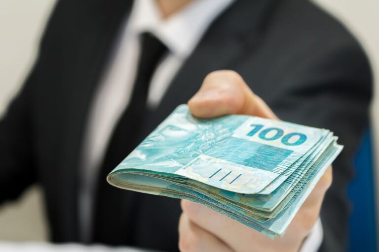 Imagem de um homem branco, de terno, com várias notas de 100 reais em uma das mãos.